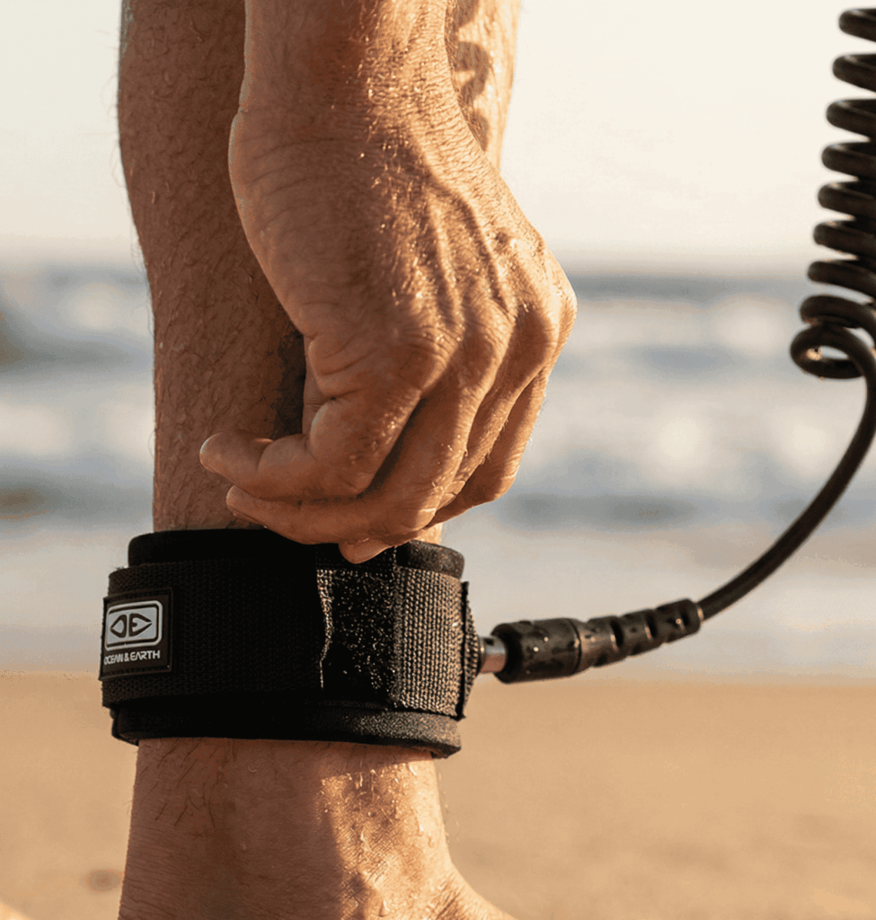 A sun-bleached hand, with visible saltwater droplets, securely fastening the velcro cuff of a new, thick (7mm), black polyurethane leash to a rugged ankle. The cuff clearly shows the brand 'Ocean & Earth'.