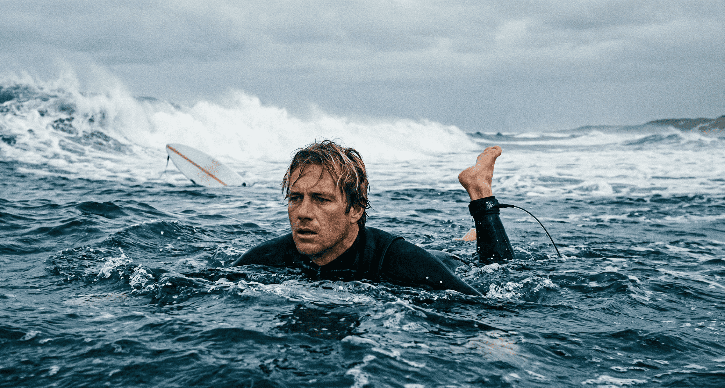 A surfer, in a 4/3mm black wetsuit with a tired, focused expression, treading water in deep, turbulent blue water past the impact zone. A thin, broken surfboard leashes string dangles from the tail plug of a white surfboard which is far in the distance, being swept shoreward by a massive, chaotic wall of whitewater.