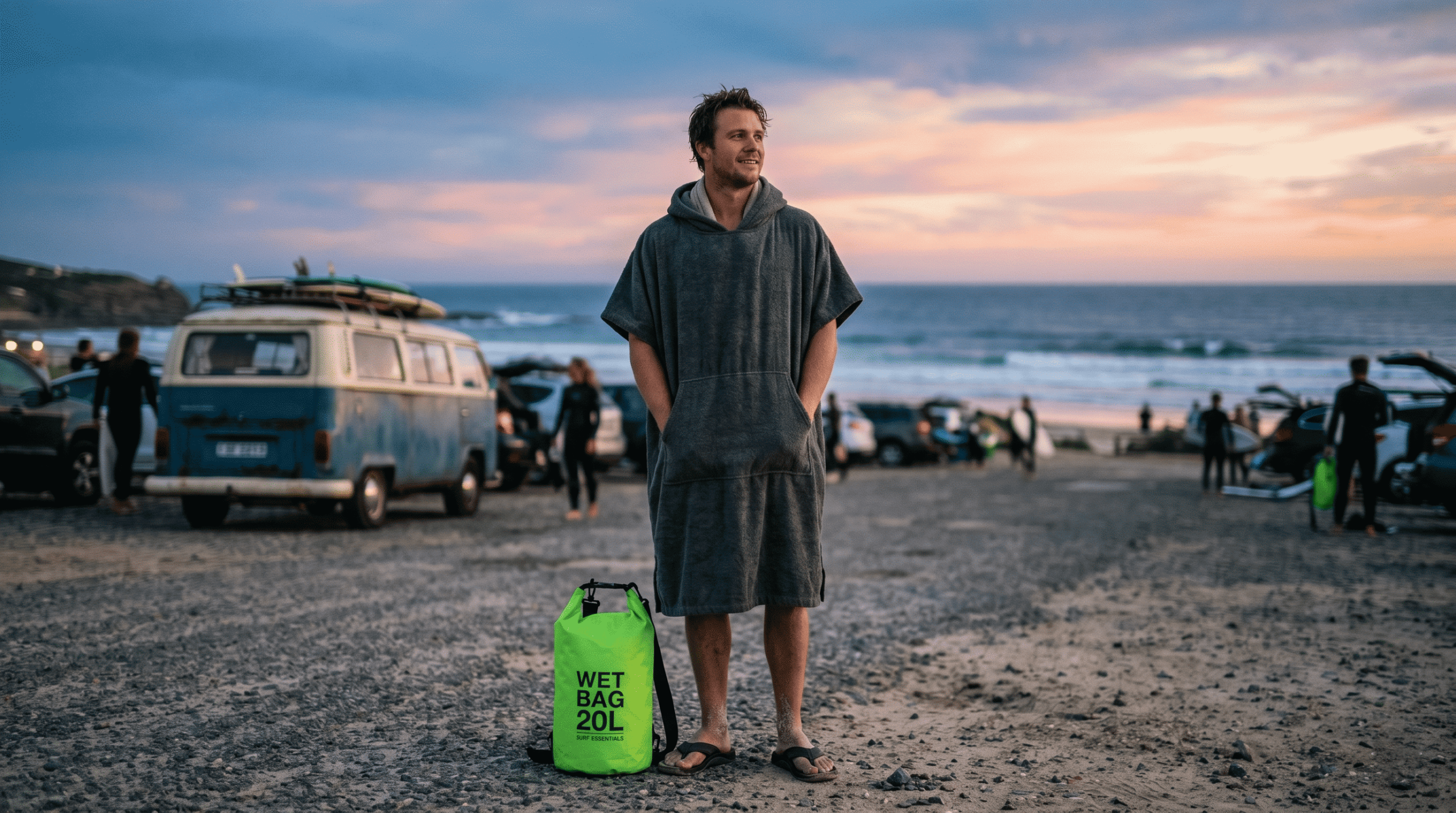 A surfer stands in a blurred beach parking lot at dusk, after a "glass-off" session. He's wearing a charcoal gray, heavyweight cotton microfiber changing poncho with an oversized hood and deep pockets.
