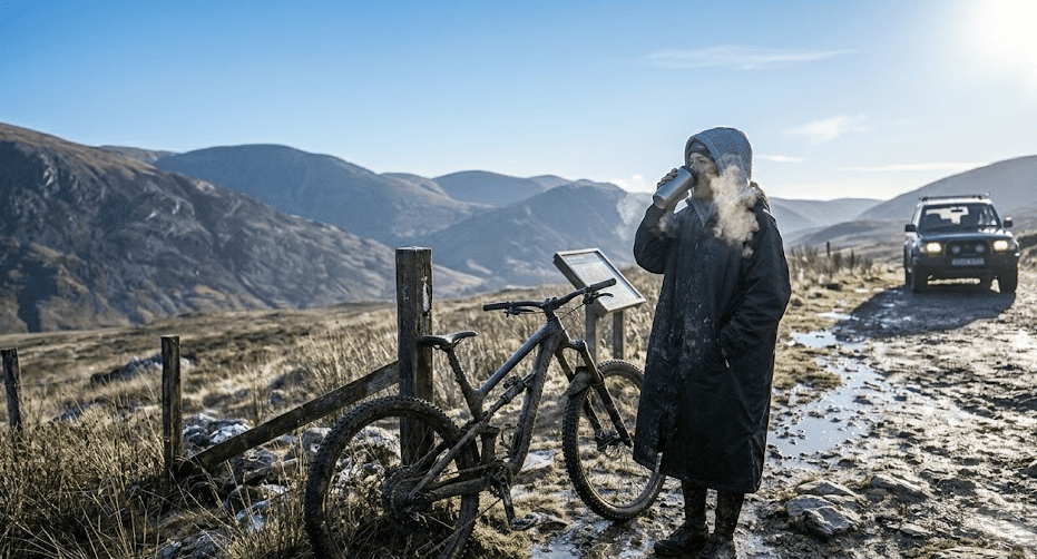 Muddy mountain biker wearing a long black weatherproof changing robe changeponcho at a windswept mountain trailhead at dusk.